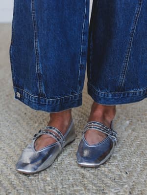 A close-up detail shot of silver metallic Mary Jane flats with double studded straps, paired with wide-leg dark wash jeans — the kind of shoe that does all the talking so your outfit doesn't have to.