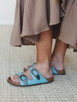 A close-up of turquoise leather double-strap sandals with engraved western buckle hardware, paired with a flowing taupe linen skirt — earthy meets unexpected in the best way.
