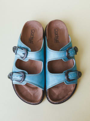 A pair of turquoise leather double-strap slide sandals shot from above against a soft sage background — the ornate silver western buckles give them that perfect mix of everyday comfort and boho personality.