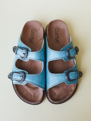A pair of turquoise leather double-strap slide sandals shot from above against a soft sage background — the ornate silver western buckles give them that perfect mix of everyday comfort and boho personality.