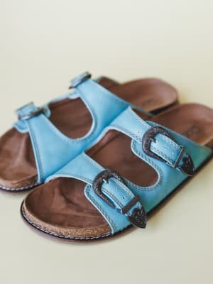 A pair of sky blue leather slide sandals with double straps, ornate western-style engraved buckles, and a worn-in brown footbed — the kind of shoes that look equally at home on a patio or a road trip.