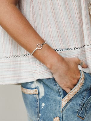A close-up lifestyle shot of a silver peace sign bangle worn on the wrist, styled casually against a striped linen top with lace trim and distressed denim — the kind of effortless everyday stack that feels intentional without trying.
