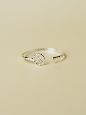 A dainty silver open-band ring with a tiny peace sign charm centerpiece, shot against a warm sand-toned backdrop — the kind of piece you put on without thinking and then never take off.