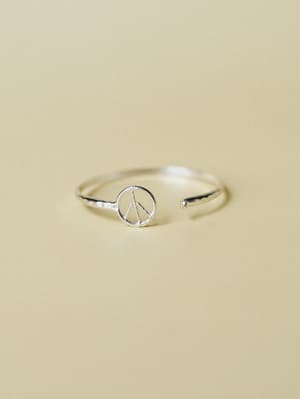 A dainty silver open-band ring with a tiny peace sign charm centerpiece, shot against a warm sand-toned backdrop — the kind of piece you put on without thinking and then never take off.
