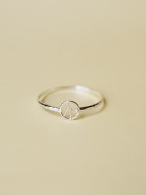 A dainty silver ring with a tiny peace sign charm sits against a soft sand-toned background — the kind of simple, meaningful piece you forget you're wearing until someone asks about it.