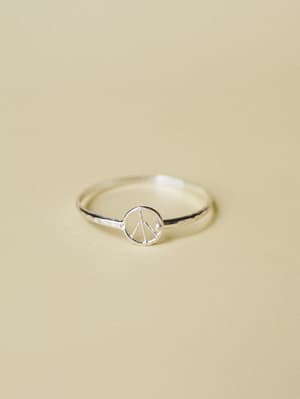 A dainty silver ring with a tiny peace sign charm sits against a soft sand-toned background — the kind of simple, meaningful piece you forget you're wearing until someone asks about it.