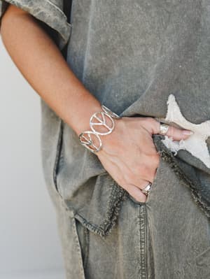 A close-up of a silver open-cuff bracelet with repeating peace sign cutouts worn on the wrist, styled against acid-washed olive denim with raw-edge trim details and stacked silver rings — effortlessly lived-in and a little free-spirited.