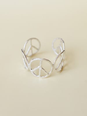 A silver open-cuff bracelet made of repeating peace sign cutouts — the kind of piece that feels equal parts vintage find and intentional statement, shot clean on a warm cream surface.