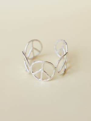 A silver open-cuff bracelet made of repeating peace sign cutouts — the kind of piece that feels equal parts vintage find and intentional statement, shot clean on a warm cream surface.
