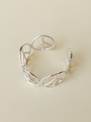 A silver open-cuff bracelet with an intertwined peace sign wire design, shot solo against a warm cream background — the kind of piece that looks like it was found at a market and worn every day since.