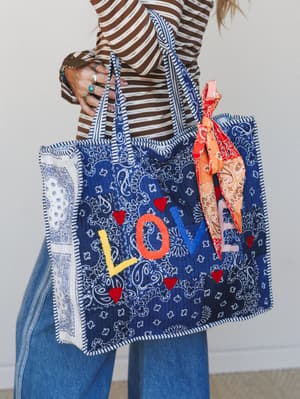 A bandana-print quilted tote bag with a colorful 'LOVE' appliqué and red heart embellishments, held by a woman in a brown striped top, wide-leg jeans, and stacked silver and turquoise rings — the kind of bag that makes you smile every time you pick i