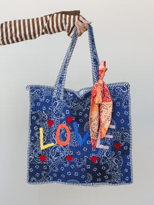 A hand holds up a quilted navy bandana-print tote bag with multicolor 'LOVE' embroidery and red heart appliqués, styled with an orange paisley scarf tied to the strap — the kind of bag that makes people ask where you got it.