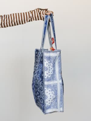A patchwork bandana-print tote held up against a soft grey wall by a hand peeking out of a brown striped sleeve — casual, expressive, and the kind of bag that goes everywhere without trying too hard.