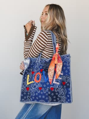 A model looks back over her shoulder showing off a large blue bandana-print quilted tote with colorful 'LOVE' appliqué lettering, red heart embellishments, and an orange paisley scarf tied to the strap — worn over a brown striped long-sleeve top and