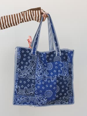 A oversized quilted tote in classic indigo bandana print held aloft by a hand in a brown striped sleeve — the kind of bag that carries your whole life and still looks good doing it.