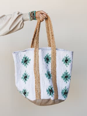 A woven tote bag in white and natural jute is held up by a model's hand stacked with beaded bracelets — the teal floral motifs and braided straps give it that easy, market-to-beach energy.