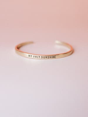 A slim gold cuff bangle stamped with 'MY ONLY SUNSHINE' rests against a soft blush gradient background — the kind of piece that feels like a little reminder you wear every day.