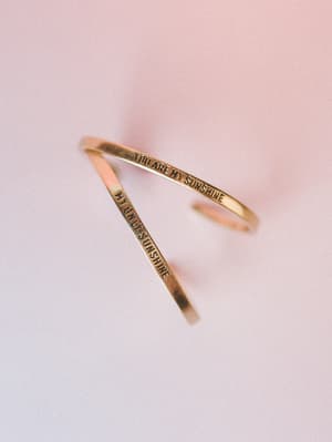 An antique gold cuff bangle engraved with 'You Are My Sunshine / My Only Sunshine' rests open on a soft blush-to-lavender gradient surface — the kind of piece that feels like a gift you keep for yourself.