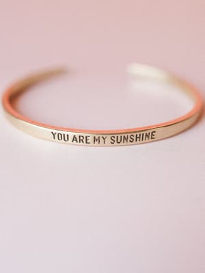 A slim gold cuff bracelet stamped with 'YOU ARE MY SUNSHINE' rests against a soft blush pink background — the kind of piece that feels like a little note from someone who loves you.