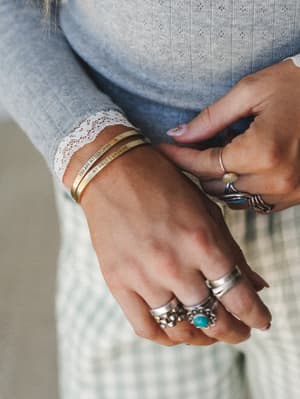 A close-up of stacked hands showing a gold engraved cuff bangle reading 'You Are My Sunshine / My Only Sunshine,' layered with silver statement rings featuring turquoise stones, against a soft blue pointelle knit sleeve with lace trim.