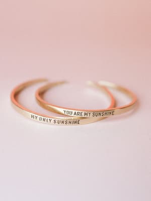 Two slim gold cuff bangles stamped with 'You Are My Sunshine' and 'My Only Sunshine' rest together against a soft blush background — the kind of jewelry that feels like a note someone meant to keep.