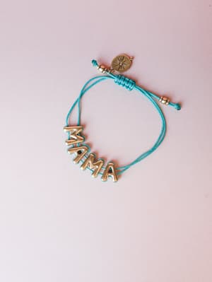 A 'MAMA' letter bracelet in antique gold on a teal adjustable cord, photographed flat against a soft blush background — the kind of piece that feels personal the moment you put it on.