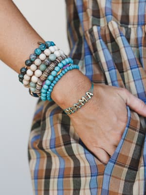 A close-up of a stacked bracelet wrist shot — the hero piece is a turquoise cord 'MAMA' letter bracelet in antique gold, layered with an earthy mix of stone, wood, and turquoise bead bracelets against a plaid shirt backdrop.