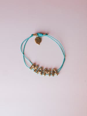 A turquoise cord bracelet with chunky antique gold 'MAMA' letter charms and a small branded coin pendant, laid flat against a soft blush background — the kind of thing you'd never want to take off.