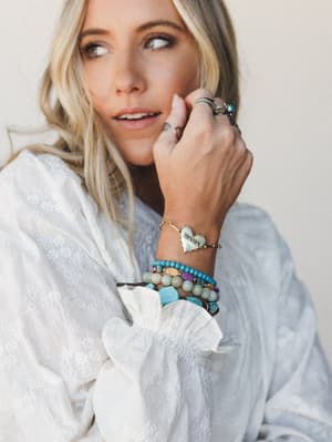 A close-up lifestyle shot of a stacked bracelet arm party on a model wearing a white floral-embroidered blouse — the star is a gold heart charm bracelet engraved with 'MAMA', layered with turquoise beaded, mixed stone, and leather bracelets, finished