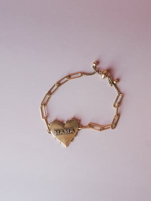 A gold paperclip chain bracelet with an engraved 'MAMA' heart charm at center, laid flat against a soft blush background — the kind of piece that lives on your wrist and never comes off.