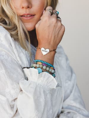 A close-up wrist shot showing a stacked bracelet look anchored by a gold chain 'MAMA' heart charm bracelet, layered with turquoise, sage, and multicolor beaded bracelets — worn with a white floral embroidered ruffle-sleeve top and a stack of silver r