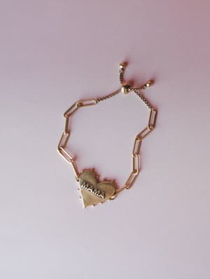 A gold paperclip chain bracelet with a spiked heart charm stamped with 'MAMA' and an adjustable sliding closure, laid flat against a soft blush-pink background — the kind of piece you wear every day and never take off.
