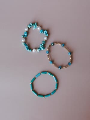 Three beaded stretch bracelets laid out on a soft mauve background — one with irregular turquoise chips and pearl accents, one with gold ball beads and faceted blue stones, and one with cylindrical turquoise tubes separated by gold beads.