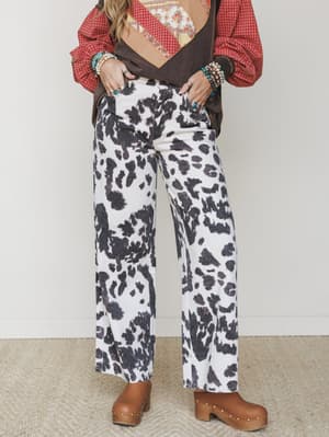 Wide-leg cow print pants styled with a layered patchwork vest, red plaid shirt, stacked turquoise jewelry, and chunky clogs — a full-on western boho moment that commits completely and somehow makes it work.