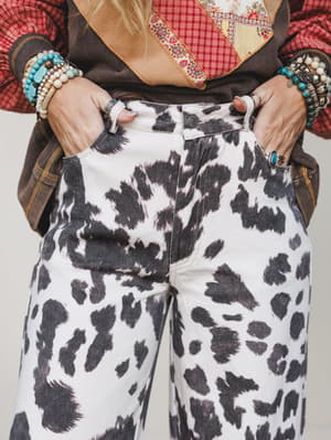 A close-cropped shot of cow print wide-leg jeans styled with a patchwork vest, plaid layer, and stacked turquoise bracelets — the kind of outfit that commits fully and doesn't apologize for it.
