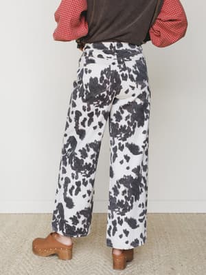 A back-view shot of wide-leg cow print pants styled with a red gingham shirt, dark vest, and tan wood-platform clogs — earthy, expressive, and a little bit ranch-meets-vintage.