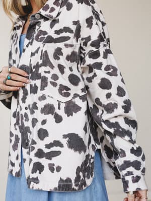 A close-up torso shot of an oversized cow print shacket layered over a light blue dress, styled with stacked silver and turquoise rings — the kind of piece that feels effortlessly collected without trying too hard.