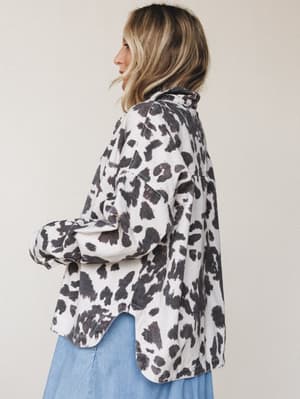 A relaxed, oversized cow print jacket shot from behind at a three-quarter angle — worn over a light blue midi skirt, the contrast between the bold animal print and soft denim feels effortlessly put-together without trying too hard.