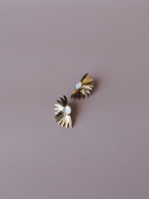 A pair of gold sunburst stud earrings with oval moonstone centers rest casually against a muted mauve backdrop — the kind of earrings that feel like they've always been yours.