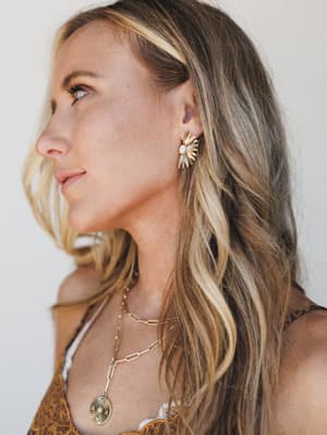A close profile shot that lets the gold sunburst stud earring do its thing — warm, glowy, and effortlessly layered with chain necklaces against a soft neutral backdrop.