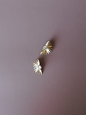 A pair of gold sunburst stud earrings with pearl centers rest casually on a muted mauve surface — the kind of earrings you reach for when you want the outfit to feel intentional without trying too hard.
