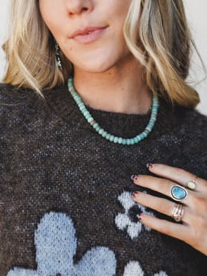 A close-up lifestyle shot showing a turquoise beaded necklace layered over a charcoal floral knit sweater, styled with silver stone rings and dangling earrings — the kind of effortless accessory stack that looks collected, not coordinated.