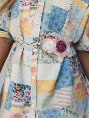 A close-up detail shot of a patchwork print shirt dress cinched with a braided fabric belt adorned with handcrafted rosette flowers in blush and dusty plum — the kind of piece that looks like it came from someone's most beautiful vintage collection.