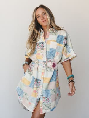 A patchwork floral shirt dress belted at the waist with a braided natural rope belt accented by fabric roses — the whole look has that 'found it at a vintage market and it fits perfectly' energy.