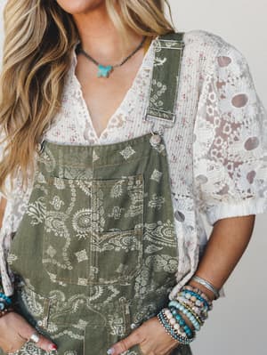 A close-up styled shot of paisley-printed olive overalls layered over a floral lace top, accessorized with stacked beaded bracelets and a turquoise butterfly necklace — the kind of outfit that looks like it came together effortlessly but absolutely d
