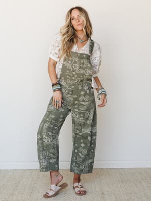 A relaxed full-body shot of a model wearing olive paisley-print wide-leg overalls layered over a white lace top, styled with stacked beaded bracelets and flat sandals — effortless boho energy with a vintage-western nod.