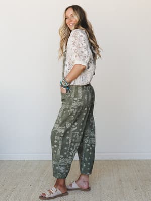 A smiling model shot in profile shows off a relaxed, lived-in pairing of olive bandana-print overalls with a sheer white lace top — the kind of effortless boho outfit that somehow looks both thrown-together and totally intentional.