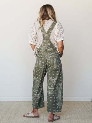 Shot from the back, a model wears wide-leg paisley print overalls in an olive army green layered over a cream eyelet lace top — the kind of outfit that looks like it came together effortlessly but was absolutely thought through.