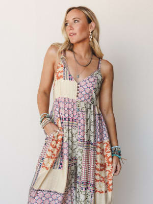 A flowy patchwork maxi dress in warm mixed prints — rust, cream, and olive — styled with layered turquoise bracelets and delicate drop earrings against a soft neutral wall.