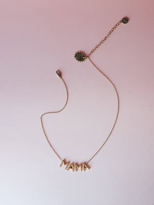 A gold 'MAMA' letter necklace laid flat against a soft pink-to-white gradient background — simple, sentimental, and the kind of thing you wear every day without taking off.